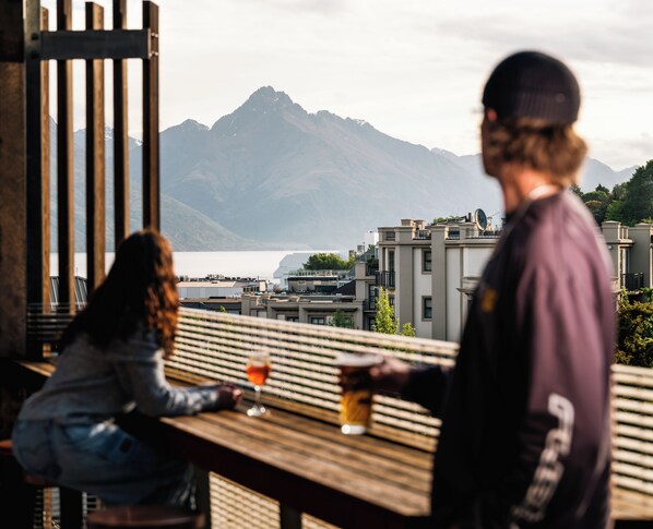 Una persona sentada a una mesa con una bebida, con vistas a un paisaje montañoso y edificios.