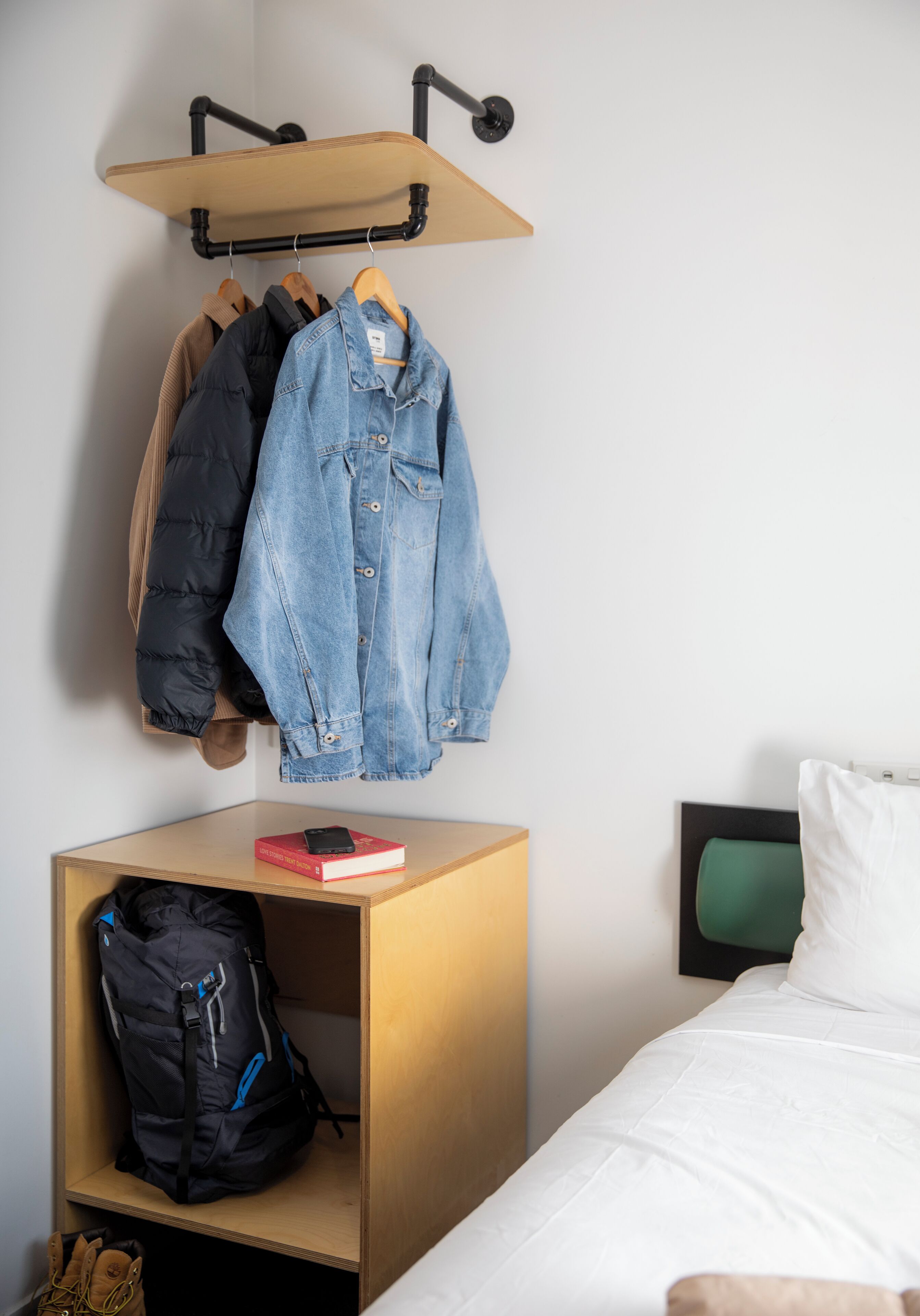 A bedroom with a bed, a bedside table with a book and a phone, a hanging clothes rack with jackets, and a small wooden shelf with a backpack.