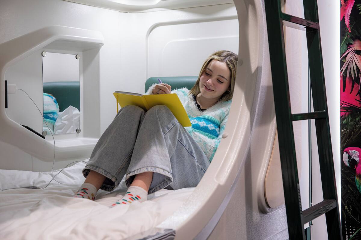 Eine Person sitzt auf einem Bett in einem Zimmer mit einem Fenster, einem Spiegel und einem kleinen Nachttisch.