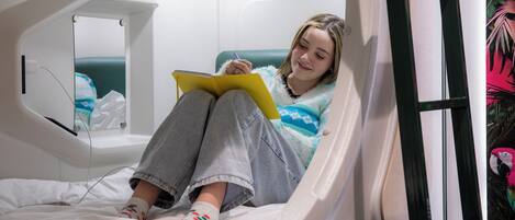 A person sitting on a bed in a room with a window, a mirror, and a small bedside table.