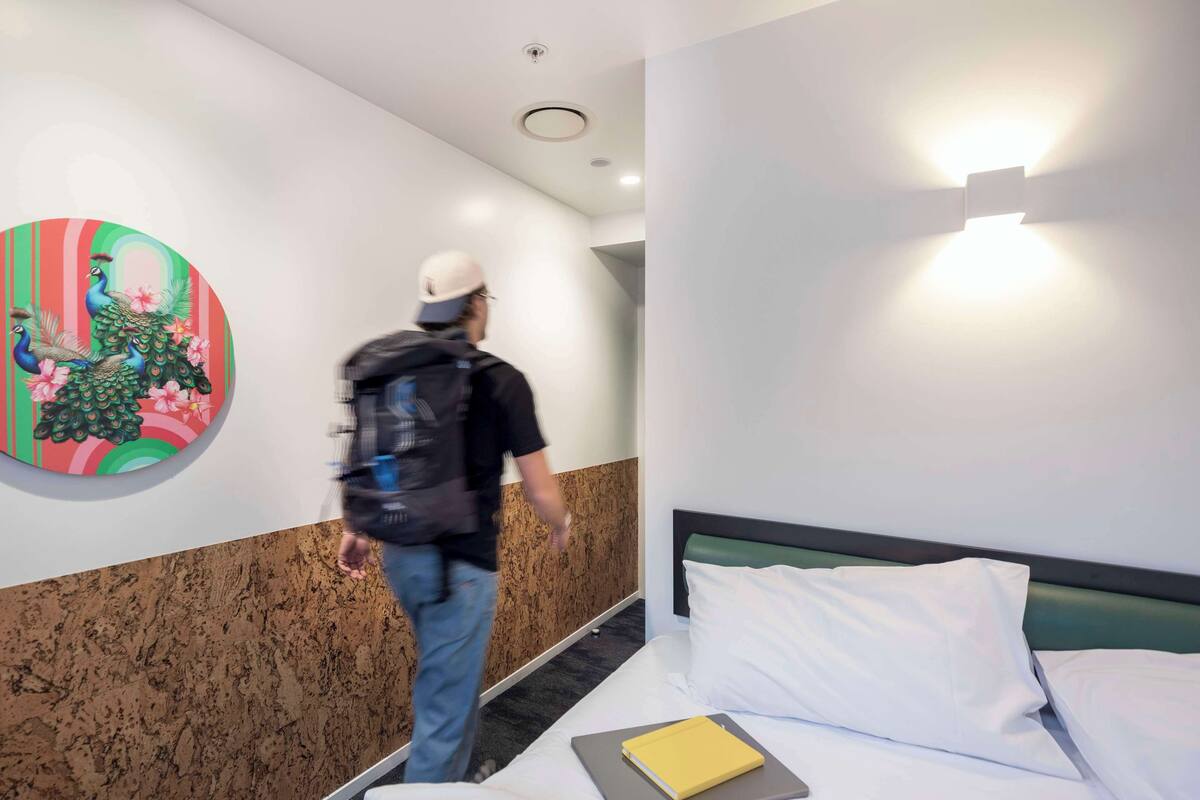 Ein Hotelzimmer mit einem Bett, einem Nachttisch mit einem Buch, einer Wand mit einem farbenfrohen Kunstwerk und einem Flur mit einer Person, die dort entlanggeht.