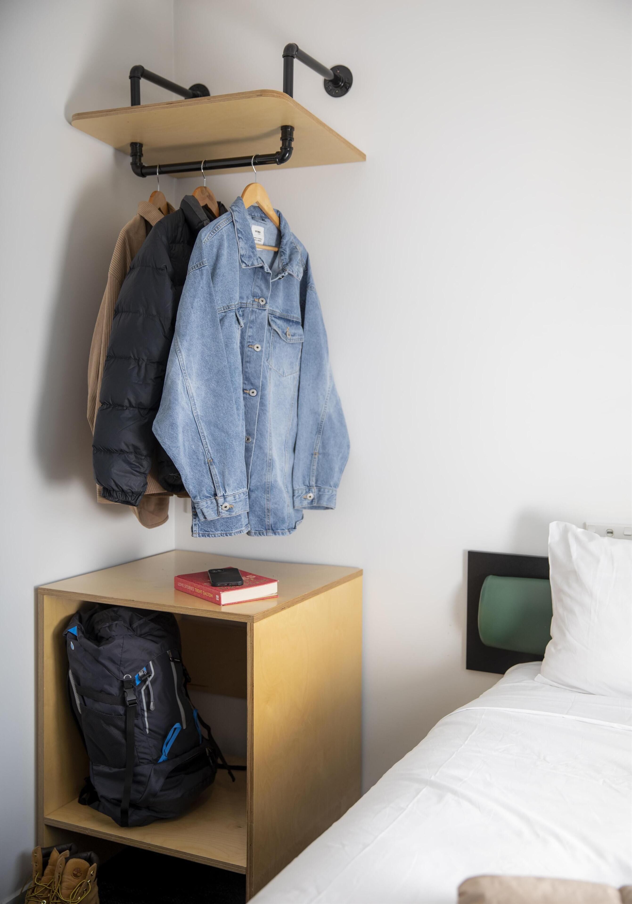 A bedroom with a bed, a bedside table with a book and a phone, a hanging clothes rack with jackets, and a small shelf with a backpack.