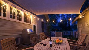 Un patio al aire libre con una mesa redonda de madera, sillas, una parrilla y luces colgantes.