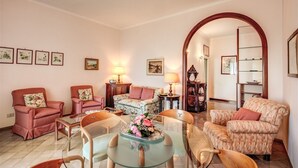 A living room with a round glass table, floral armchairs, and a sofa. There are framed pictures on the walls and a lamp on a side table.