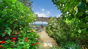 A garden pathway leading to a wooden bridge over a body of water, surrounded by lush greenery and flowering plants.