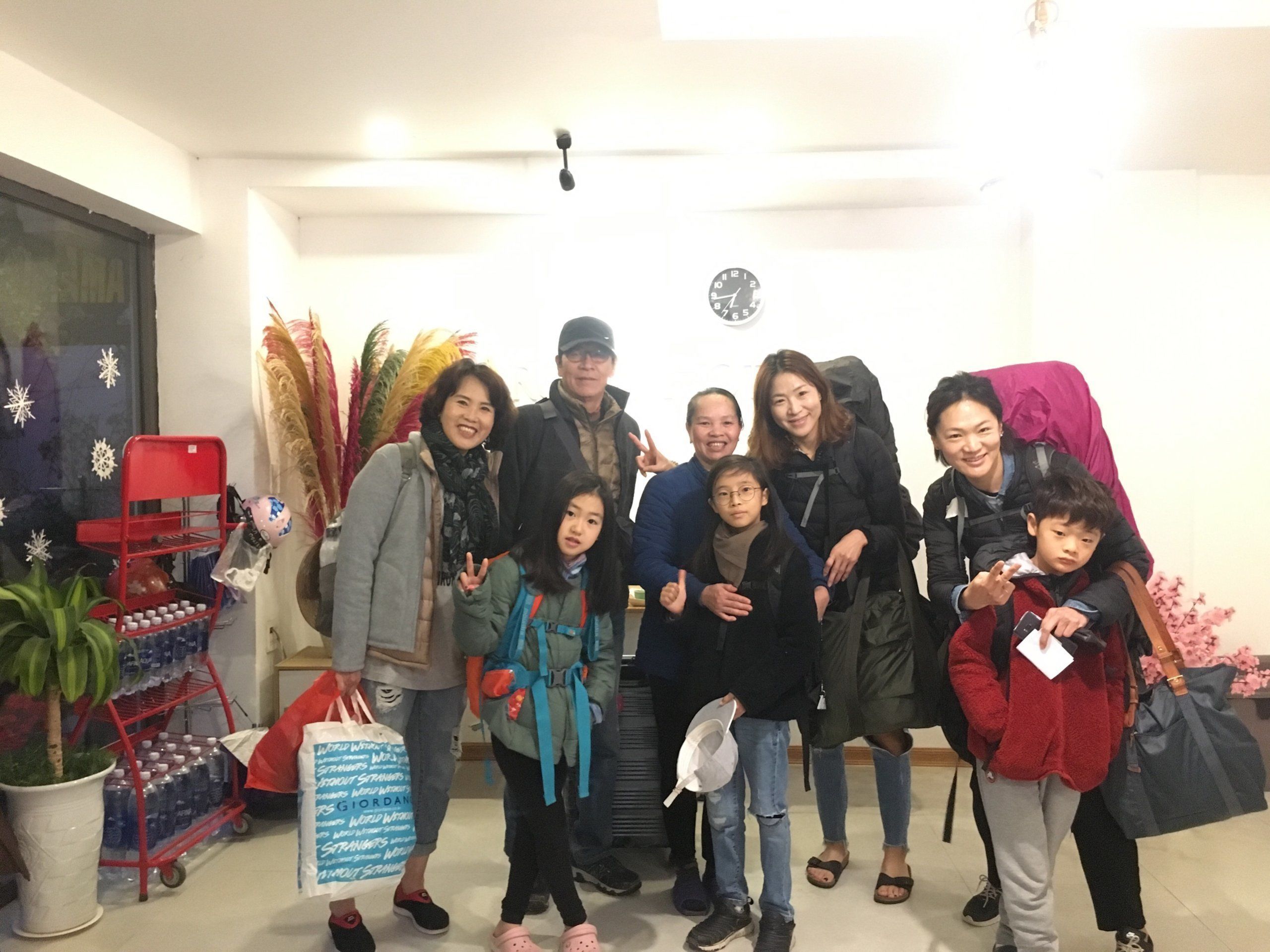 A group of people in a room with a red cart, a clock on the wall, and a potted plant.