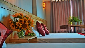 Una habitación de hotel con una cama, una mesita de noche de madera con un jarrón de girasoles, un frutero y una mesa con una planta en maceta.