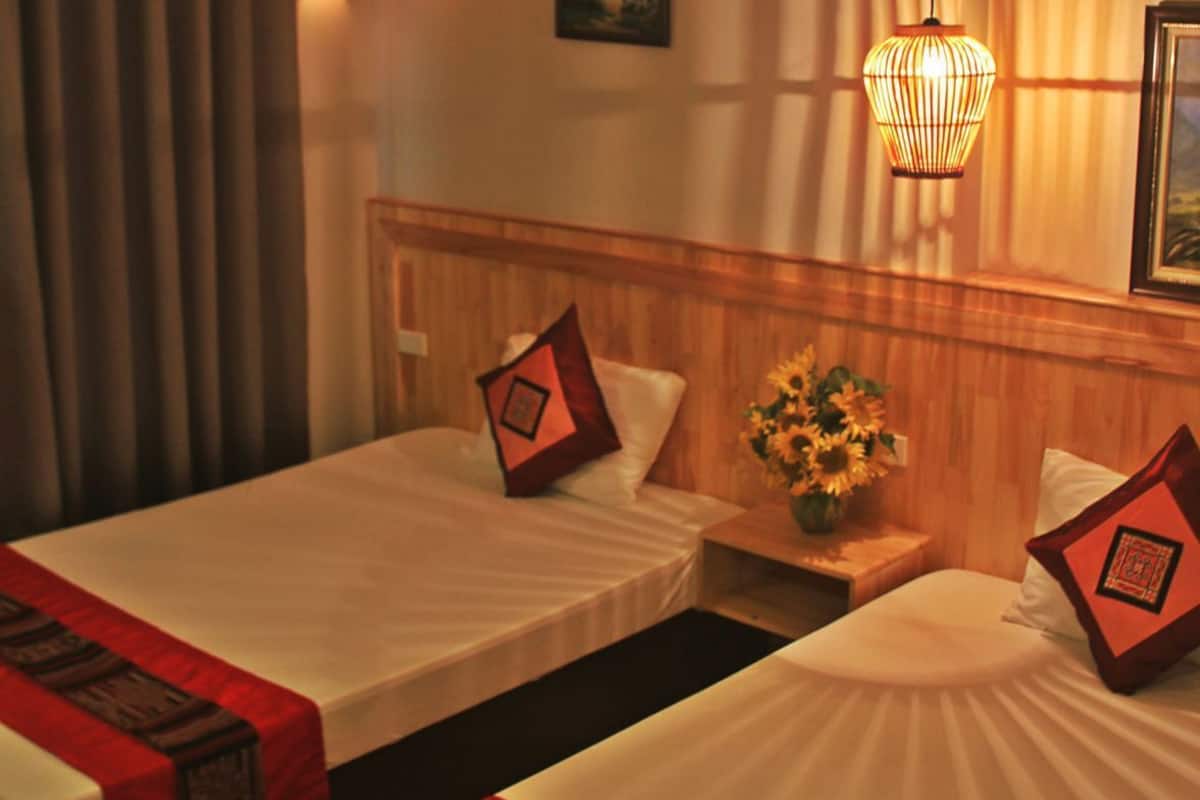 A hotel room with two beds, a nightstand with a vase of sunflowers, a wall-mounted lamp, and a painting.