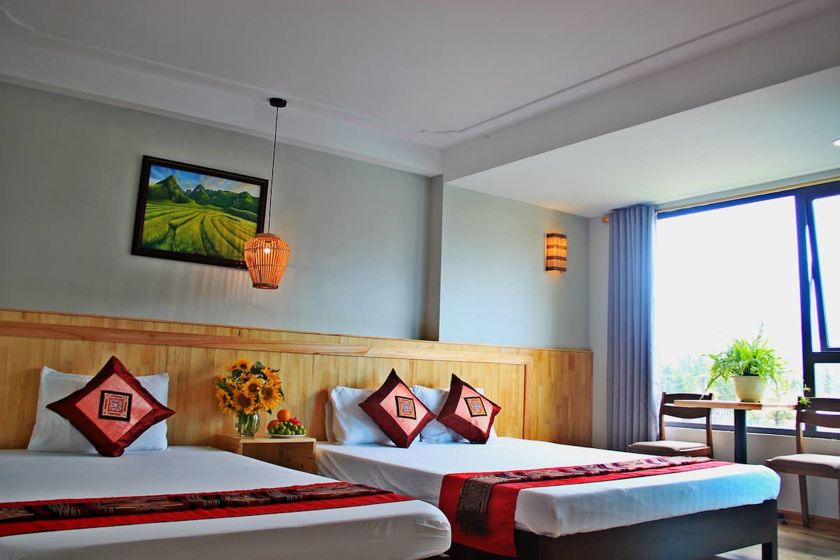 A hotel room with two beds, a wooden headboard, a painting of a landscape, a hanging lamp, a window with curtains, and a small table with a plant.