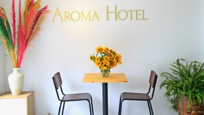 Zona de recepción de un hotel con un letrero que dice "Aroma Hotel", una mesa de madera con un jarrón de girasoles, dos sillas y una planta en maceta.