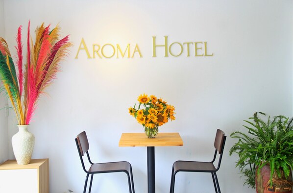 Zona de recepción de un hotel con un letrero que dice "Aroma Hotel", una mesa de madera con un jarrón de girasoles, dos sillas y una planta en maceta.