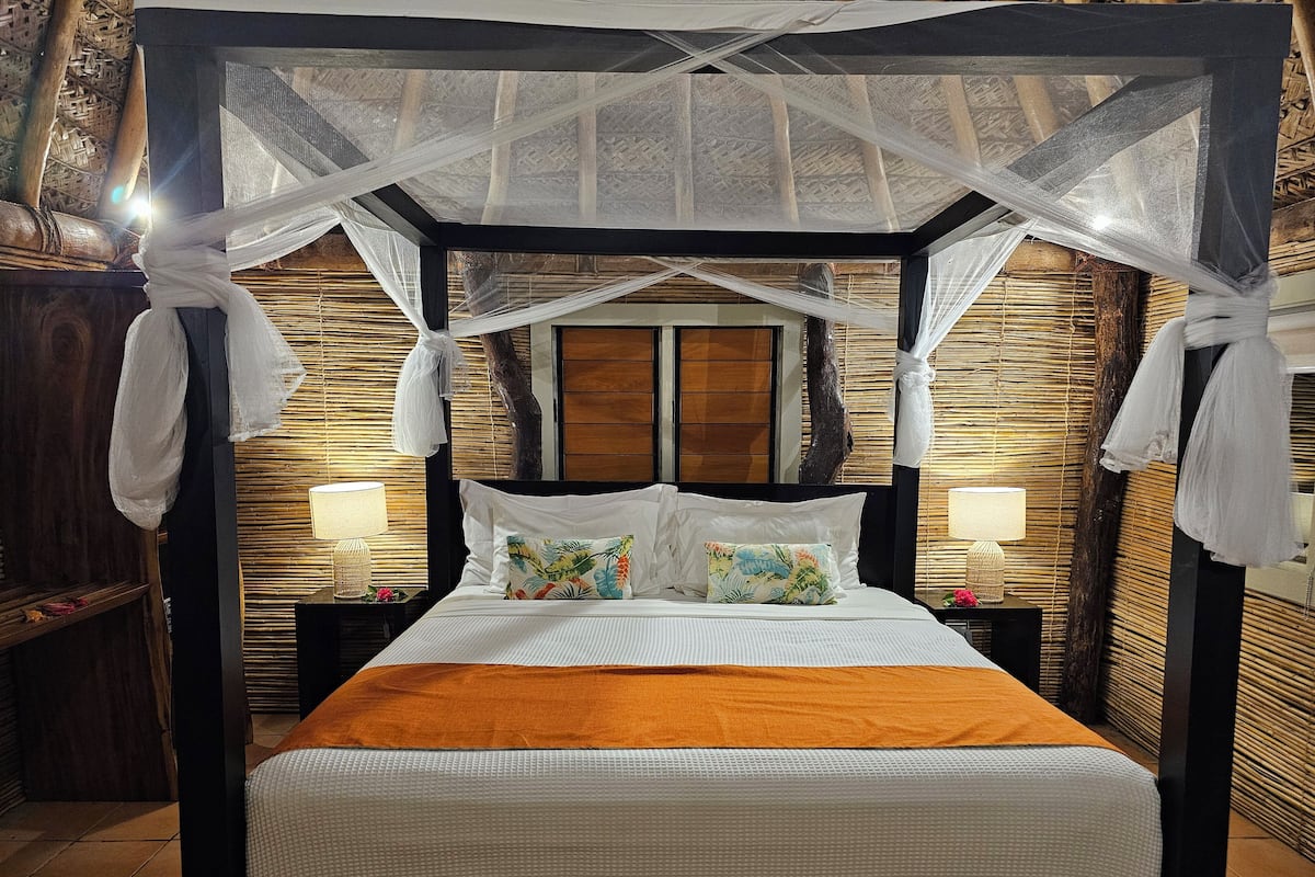 Uma cama com dossel, duas mesas de cabeceira com abajures, uma cabeceira de madeira e um painel de parede trançado.
