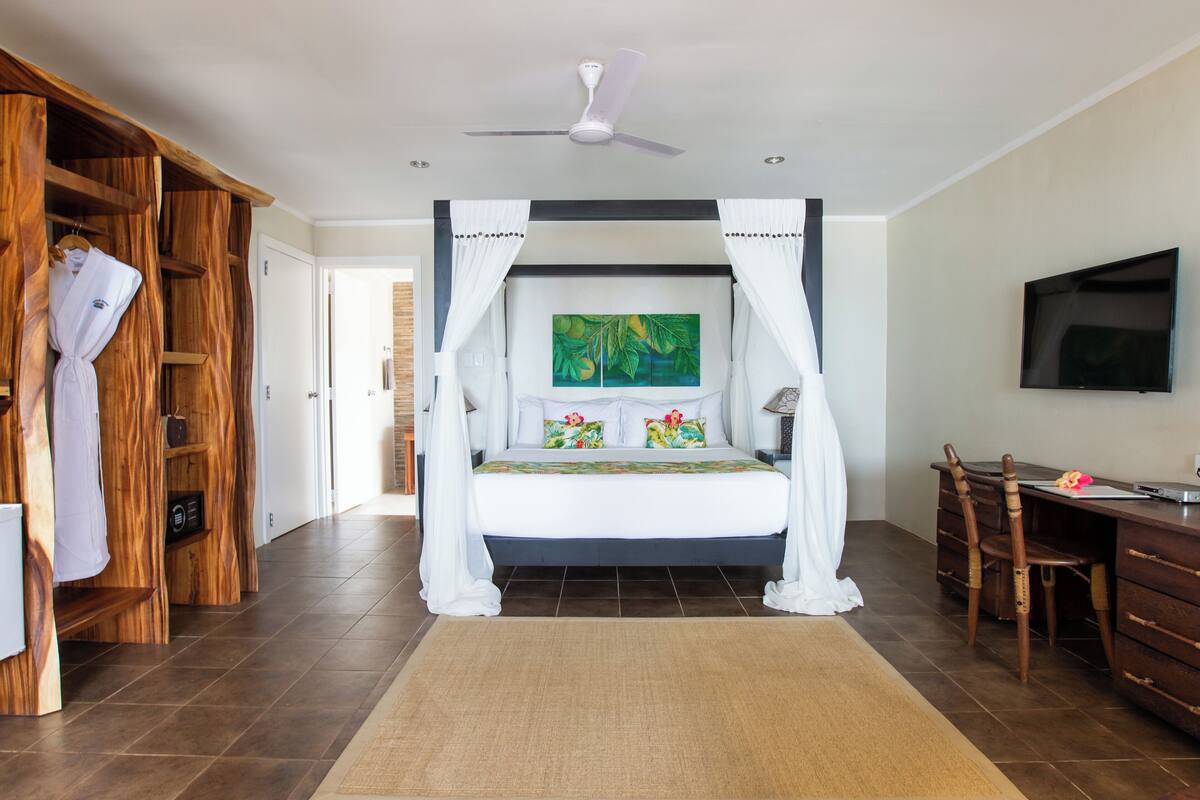 Quarto com cama de dossel, televisão montada na parede, guarda-roupa de madeira e mesa com cadeiras.