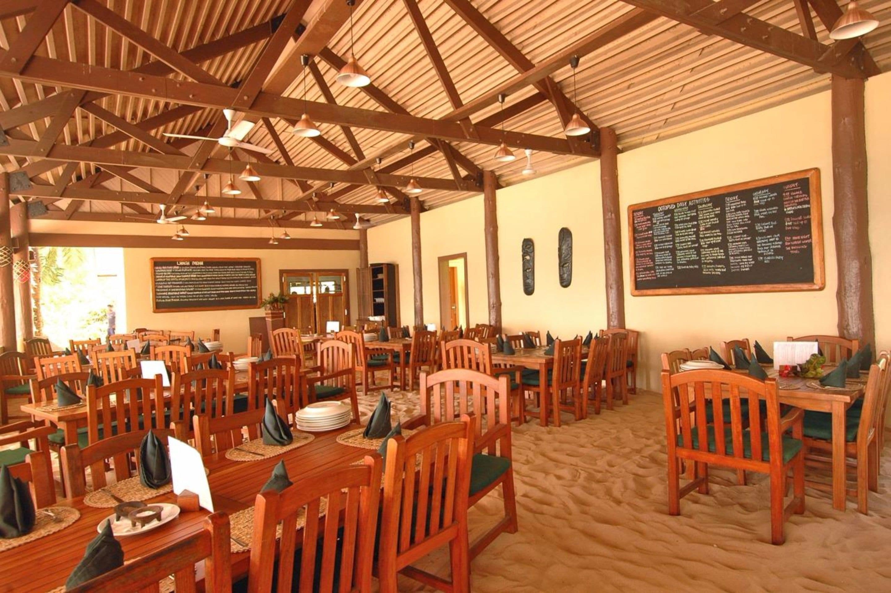 Uma sala de jantar ampla com mesas e cadeiras de madeira, teto alto com vigas aparentes e um quadro de cardápio na parede.