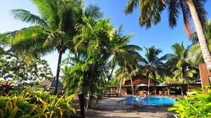 A pool area with palm trees, a wooden deck, and a building with a thatched roof in the background. - Octopus Resort (Waya Island)