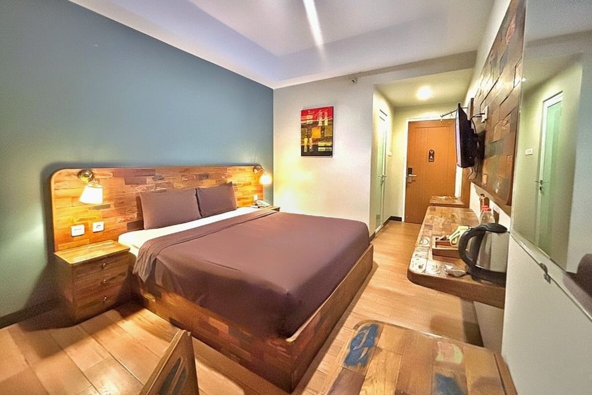Ein Schlafzimmer mit einem Holzkopfteil, einem Bett mit brauner Bettdecke, einem Nachttisch mit Lampe und einer Küchenzeile mit Kühlschrank und Mikrowelle.