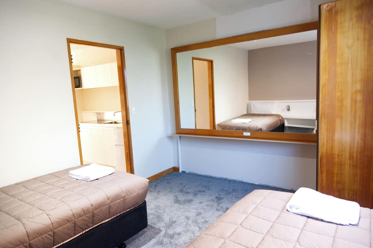 Una habitación pequeña y limpia con dos camas individuales, un espejo con marco de madera y una kitchenette al fondo.