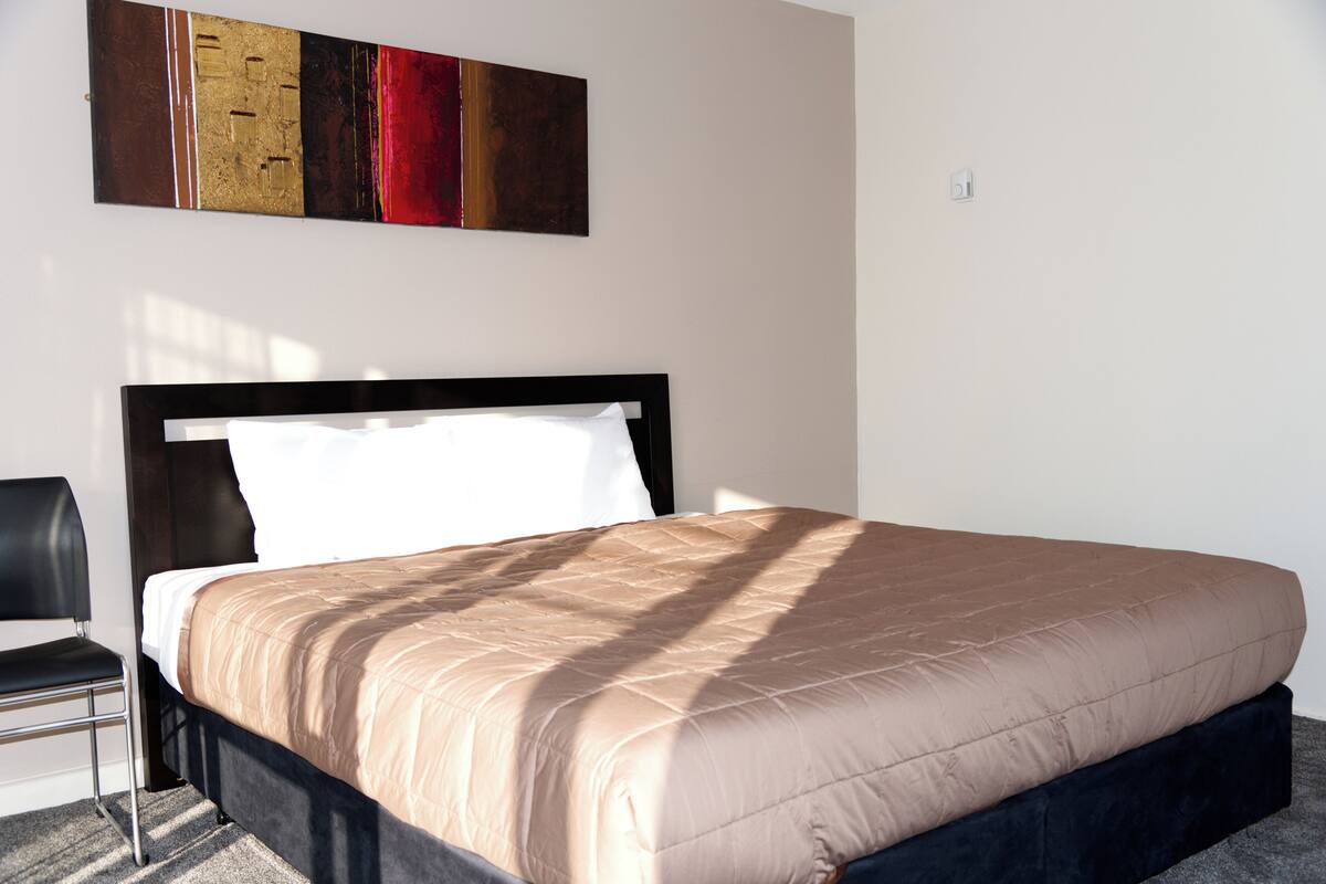 Una cama bien hecha con un cabecero negro, una silla negra y un cuadro abstracto moderno en la pared.