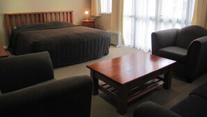 Una habitación de hotel con una cama, una mesita de noche de madera, una mesa, dos sillones y un ventanal con cortinas.