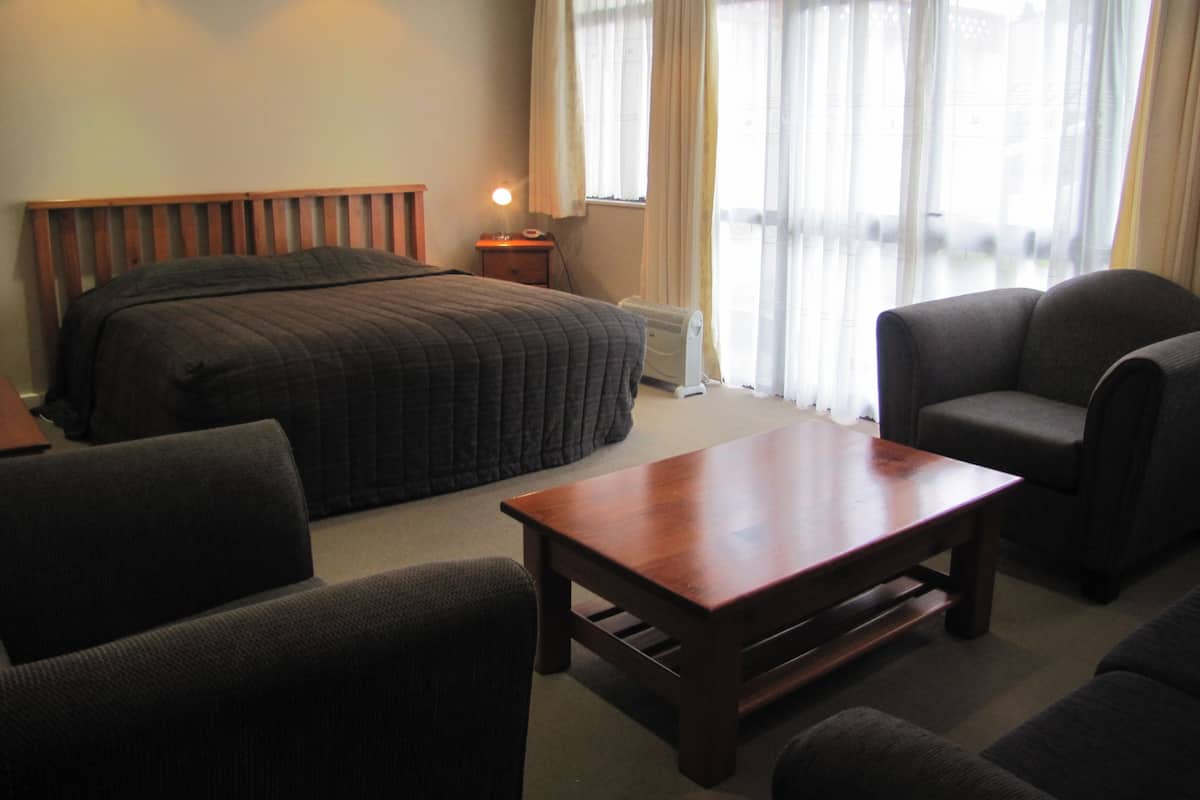 Una habitación de hotel con una cama, una mesita de noche de madera, una mesa, dos sillones y un ventanal con cortinas.