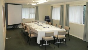 Un salón de conferencias con una mesa larga, sillas, una pantalla de proyector y ventanas con cortinas.