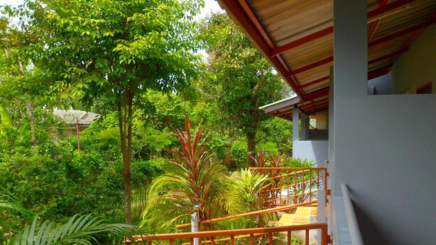 A balcony with a view of lush greenery, a wooden railing, and a building with a red roof. - Bonus Bungalow (Chalong)