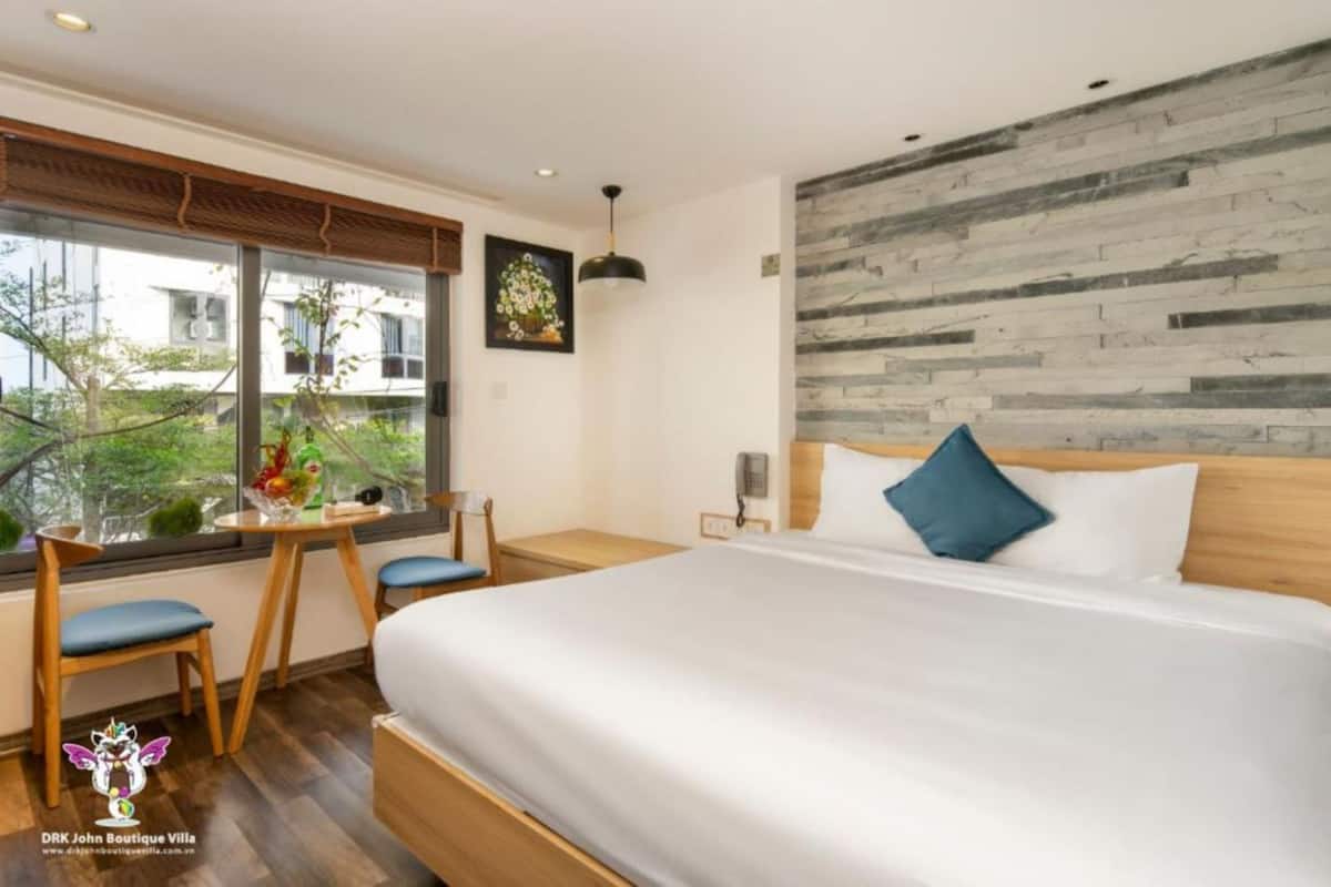 A hotel room with a large bed, a wooden desk with two chairs, a window with a view of greenery, and a wall with a textured pattern.