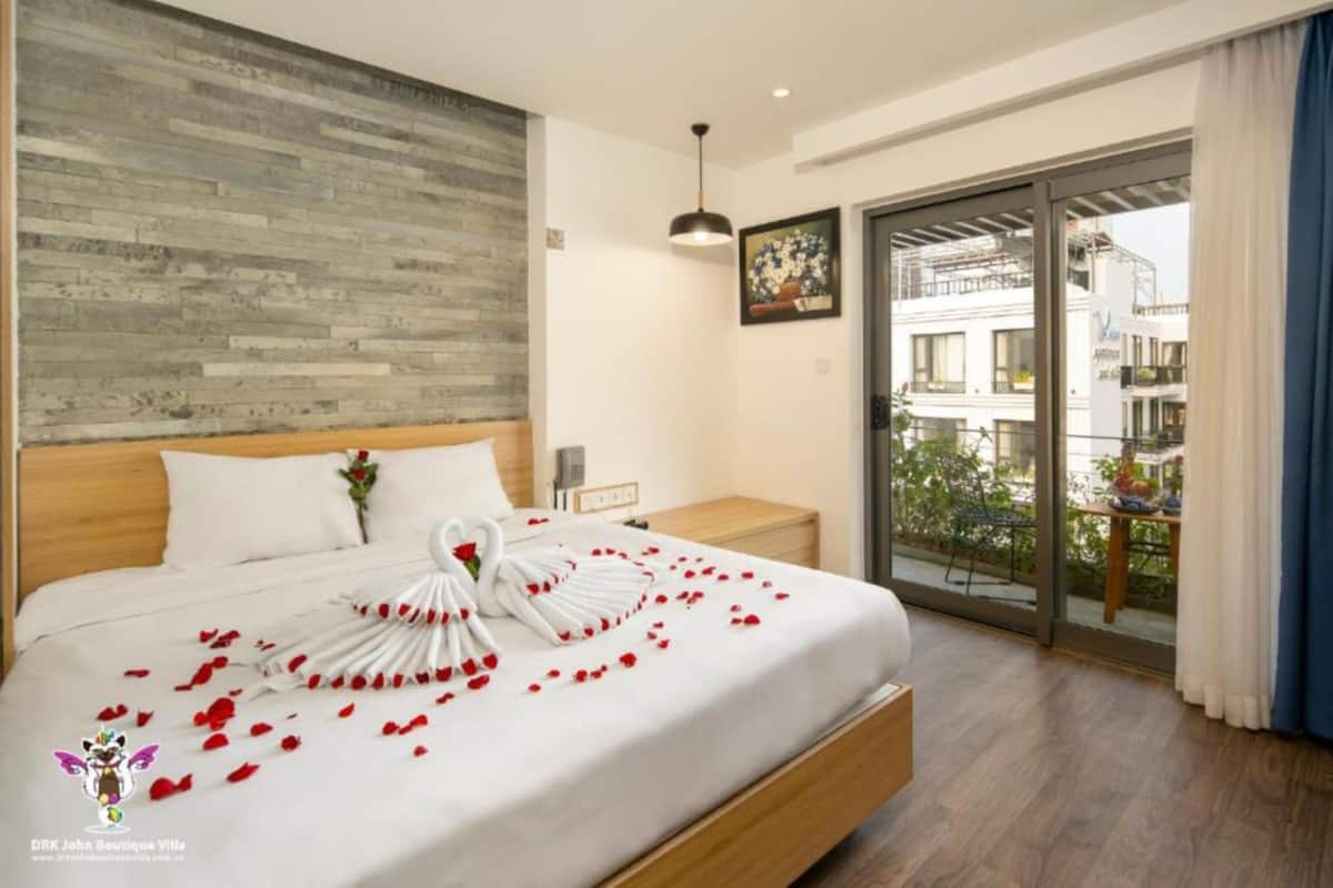A hotel room with a wooden headboard, a bed with white linens and rose petals, a nightstand, a picture frame, and a balcony with a view of buildings.