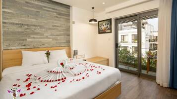 A hotel room with a wooden headboard, a bed with white linens and rose petals, a nightstand, a picture frame, and a balcony with a view of buildings.