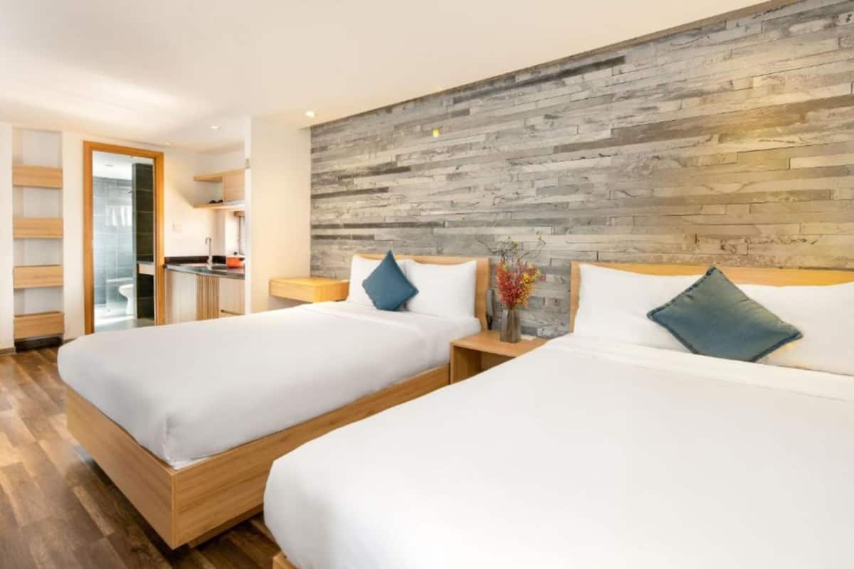 A modern hotel room with two beds, a wooden headboard, and a kitchen area visible through an open door.
