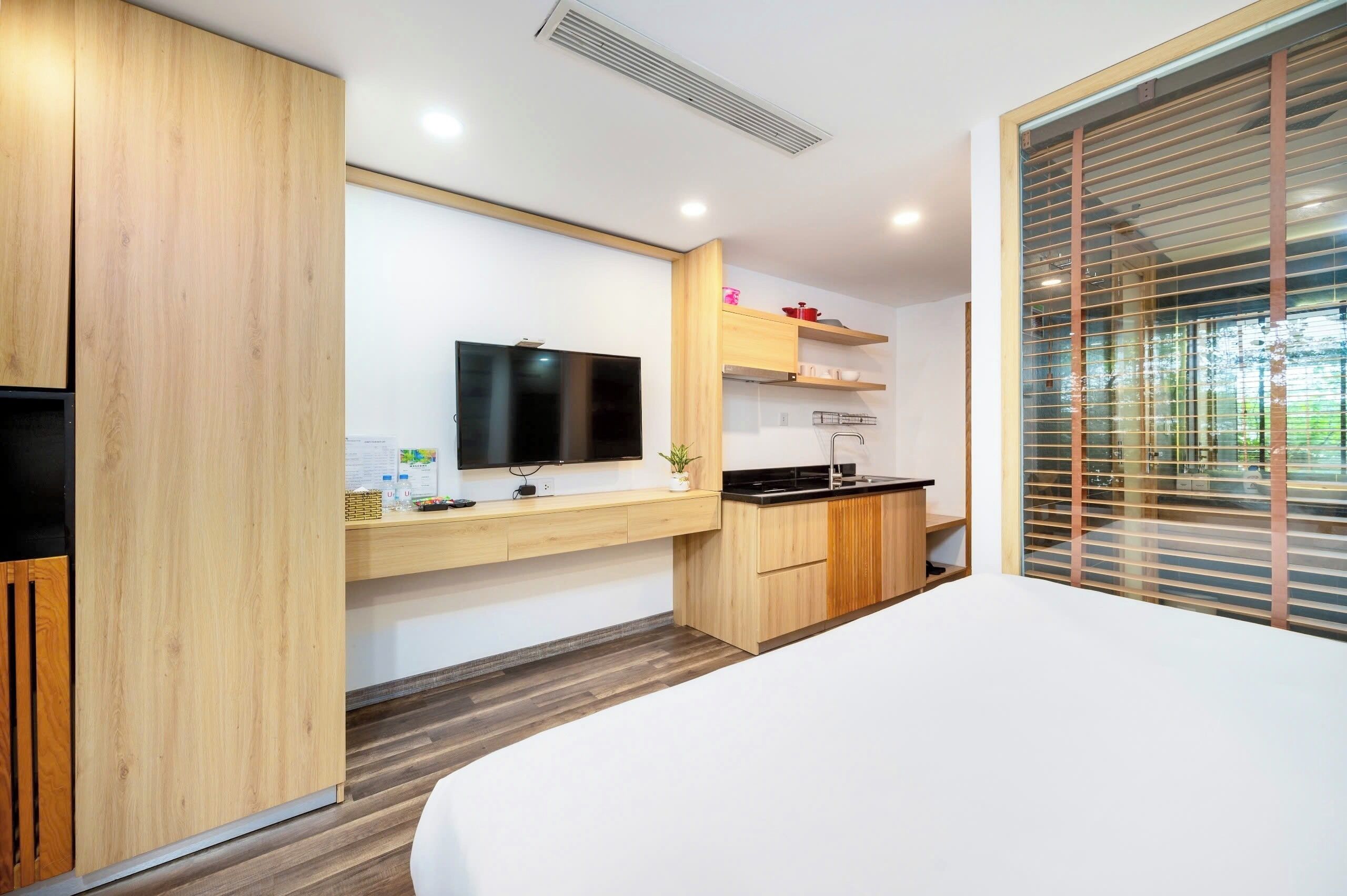 A modern hotel room with a wooden wardrobe, a flat-screen TV mounted on the wall, a small kitchen area with a stove and sink, and a large window with blinds.