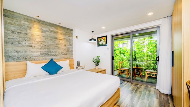 A modern bedroom with a large bed, a wooden headboard, a bedside table, a nightstand, a wall-mounted light, and a sliding glass door leading to a balcony with outdoor furniture. -  John Boutique Villa  (Da Nang)