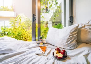A neatly made bed with a book, a plate of fruit, and a beverage in a hotel room with a view of greenery outside.