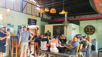 A bustling indoor dining area with hanging pendant lights, a chalkboard menu, and a fan.