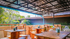 An outdoor seating area with wooden tables and benches, a covered roof, and greenery in the background. - 2 Home Resort (Chalong)