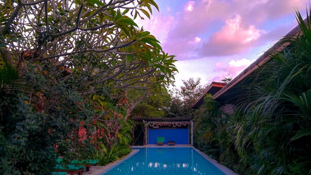 A swimming pool surrounded by lush greenery and a blue structure in the background. - 2 Home Resort (Chalong)