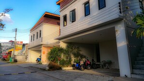 A two-story building with a flat roof and a covered entrance. There are two motorcycles parked in front, and a red truck is visible in the background. - 2 Home Resort (Chalong)