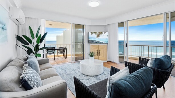 Una sala de estar moderna con sofá, mesa de centro y sillas, un balcón con vista al mar y un televisor en la pared.