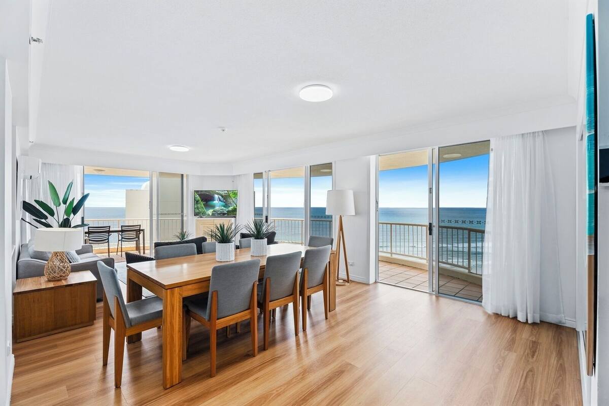 A modern living room with a wooden dining table, grey chairs, a sofa, and a TV. The room has large windows with a view of the ocean.