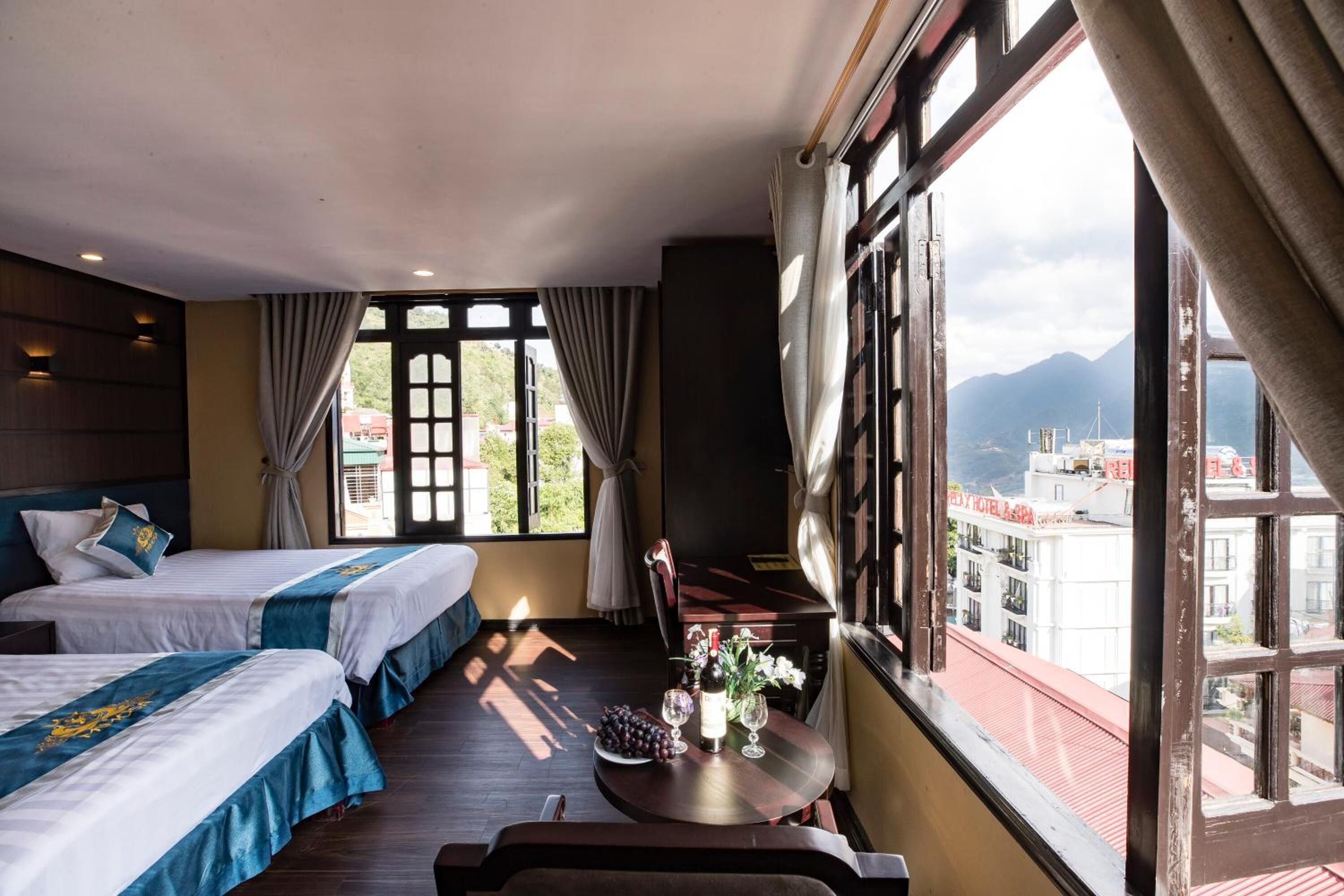 Ein Hotelzimmer mit zwei Betten, einem Esstisch mit einer Vase und Weingläsern sowie Blick auf Gebäude und Berge durch große Fenster.