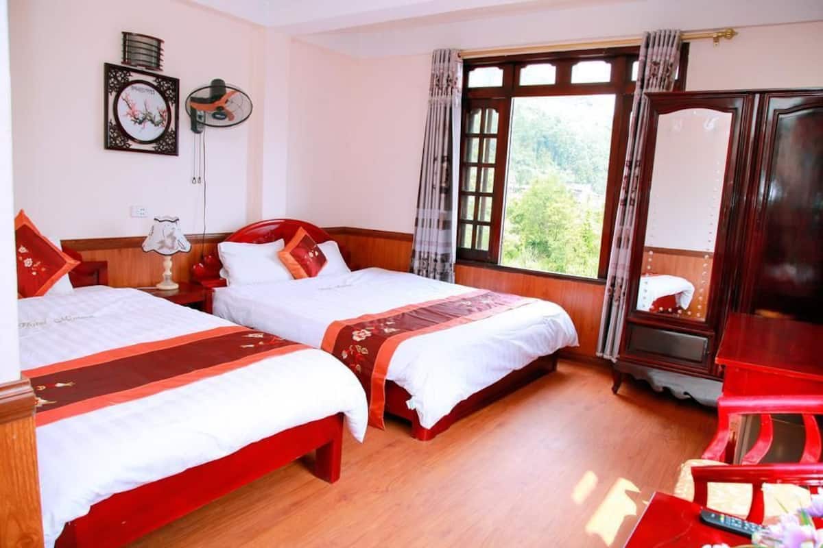 Habitación de hotel con dos camas, suelo de madera, un ventanal grande con cortinas, un armario de madera y una silla roja.