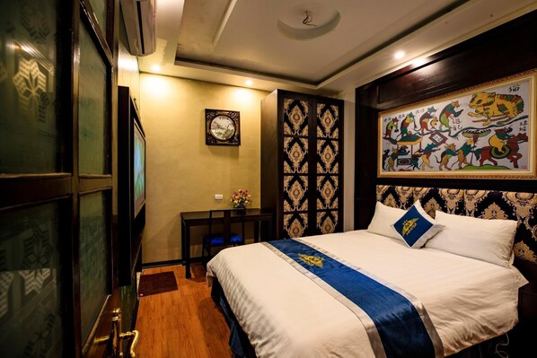 Quarto de hotel com cama, escrivaninha, cadeira, televisão fixada na parede e painel decorativo.