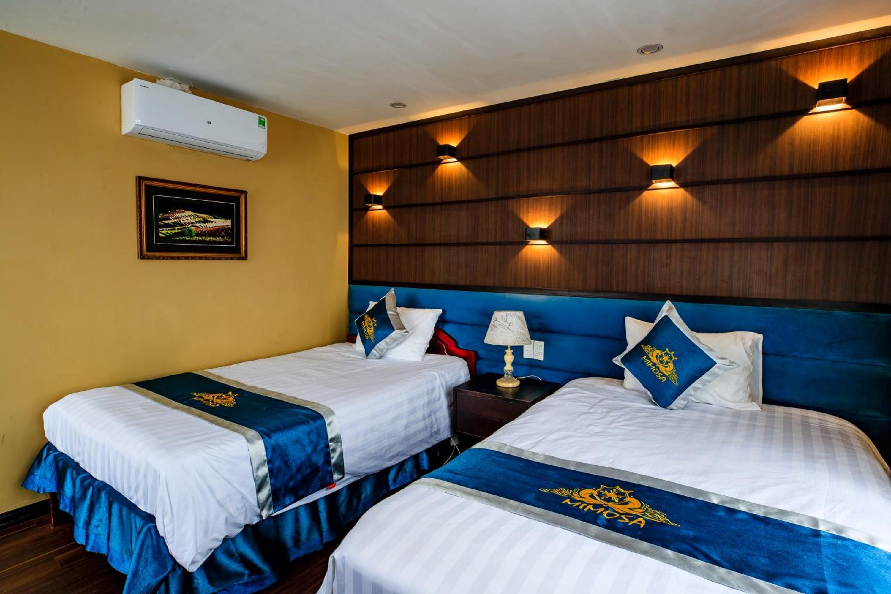 A hotel room with two beds, each with a blue bedspread and a white headboard. There is a framed picture on the wall, a nightstand with a lamp, and a wall-mounted air conditioner.
