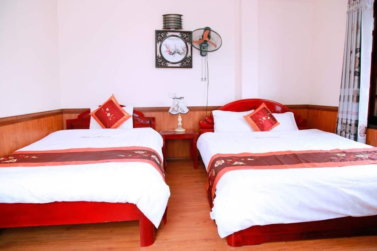 Habitación de hotel con dos camas individuales, cabeceros de madera, una mesita de noche con lámpara, un ventilador de pared y un cuadro enmarcado.