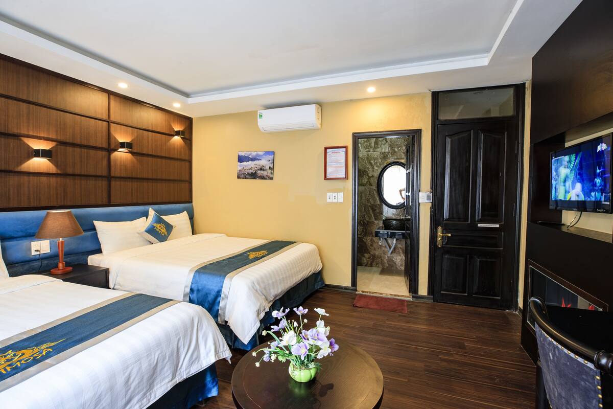 Habitación de hotel con dos camas, un televisor, aire acondicionado, baño con espejo y una mesita con flores.