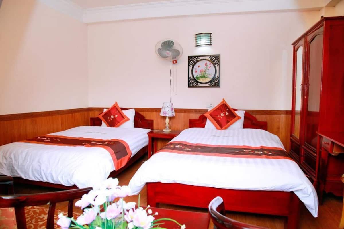 Habitación de hotel con dos camas, suelo de madera, ventilador de pared, un cuadro decorativo y un armario rojo.