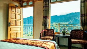 A hotel room with a bed, two chairs, a table with flowers, and a view of mountains through large windows. - Sapa Mimosa Hotel (Sa Pa)