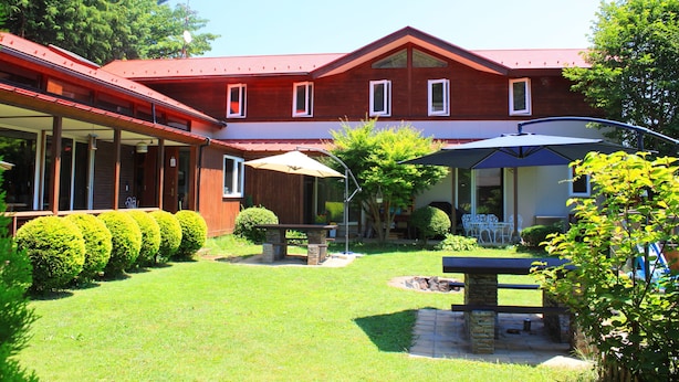 A red building with a covered patio area, a stone bench, and neatly trimmed bushes. - Pension Field (Hokuto)