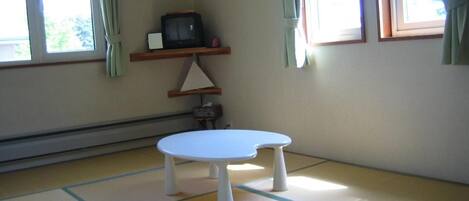 Una habitación con suelo de esteras de tatami, una pequeña mesa blanca, un televisor sobre una estantería y una ventana con cortinas.