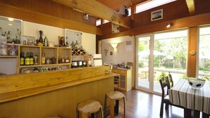 A wooden interior with a bar area, wooden stools, a table with a striped tablecloth, and a view through a glass door to an outdoor area. - Pension Field (Hokuto)